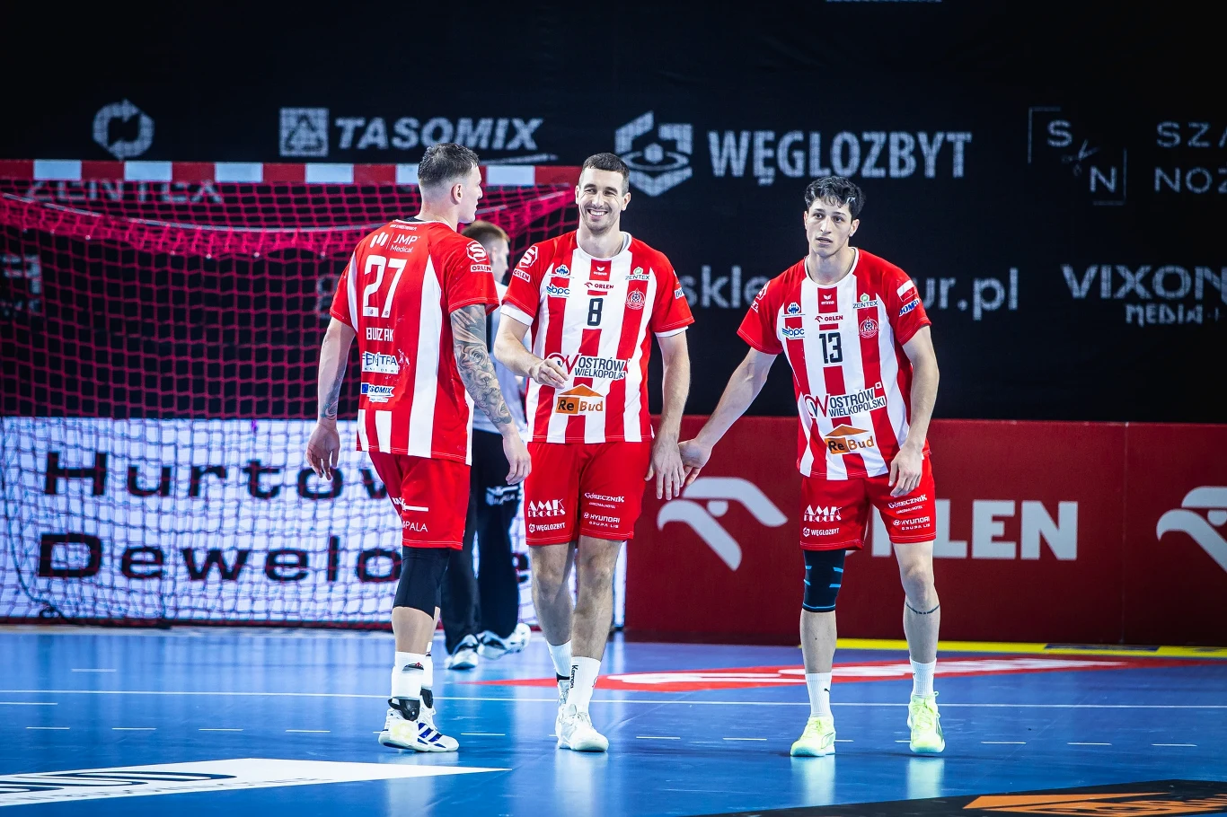 Trzej zawodnicy drużyny piłki ręcznej w czerwono-białych strojach stoją na hali sportowej, uśmiechając się i rozmawiając ze sobą, w tle bramka i reklamy sponsorów