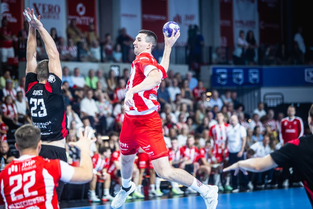 Rebud KPR Ostrovia Zawodnik w biało-czerwonym stroju wyskakuje z piłką w ręce na tle hali sportowej podczas meczu piłki ręcznej, naprzeciw niego przeciwnik w czarnym stroju próbujący zablokować rzut; w tle widoczna widownia i inni zawodnicy.