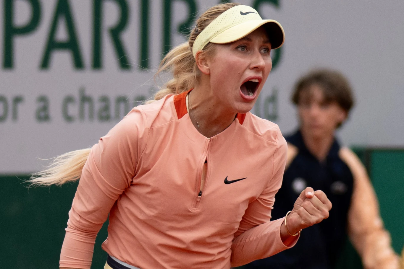 Zawodniczka tenisowa wyrażająca silne emocje podczas meczu, ubrana w sportową odzież, zaciska pięść w geście triumfu, w tle niewyraźna postać i fragment reklamy.