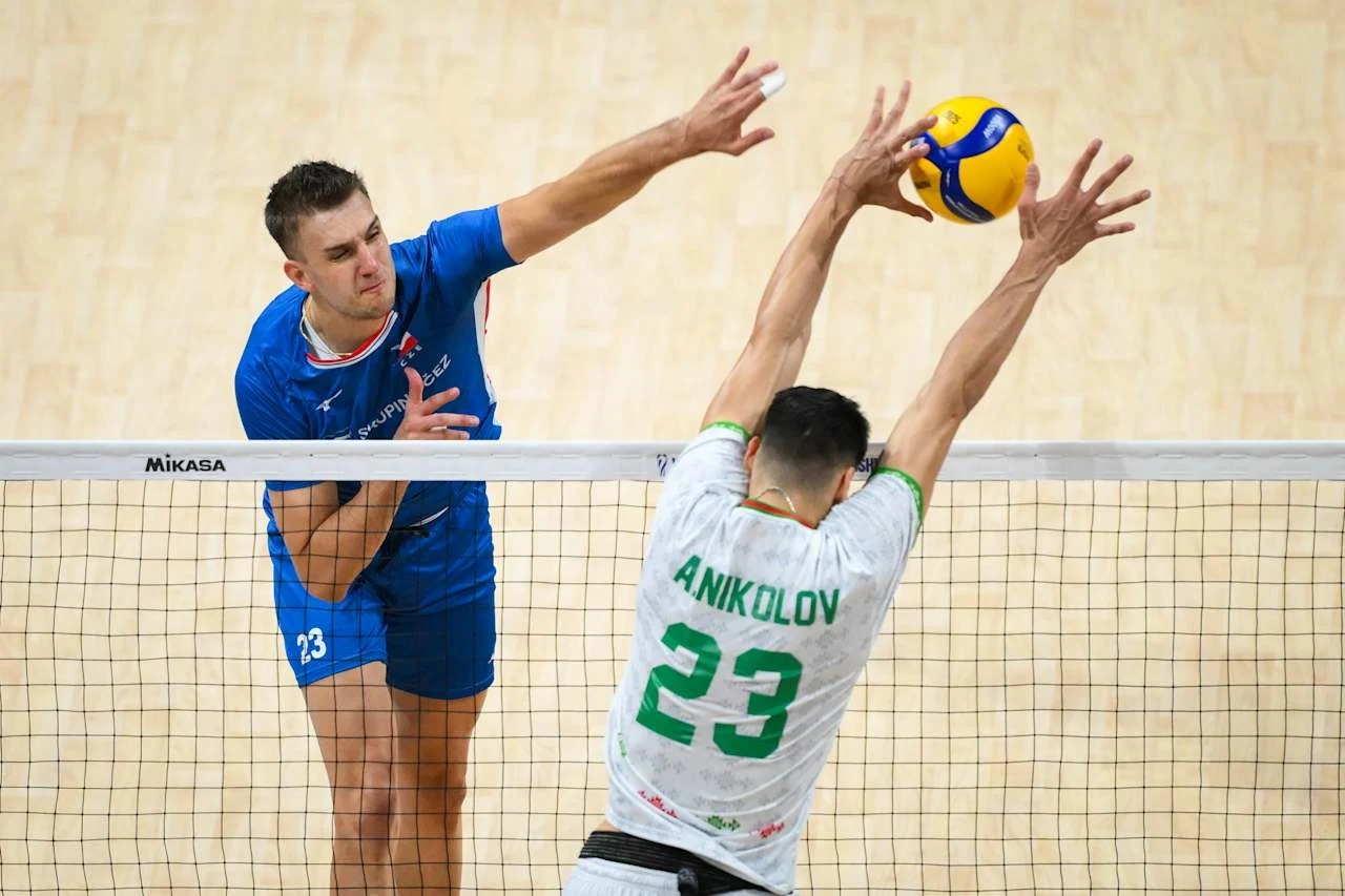 Dwóch siatkarzy w trakcie gry, jeden z zawodników w niebieskim stroju wykonuje atak nad siatką, podczas gdy drugi w białej koszulce z numerem 23 próbuje zablokować piłkę po drugiej stronie boiska.