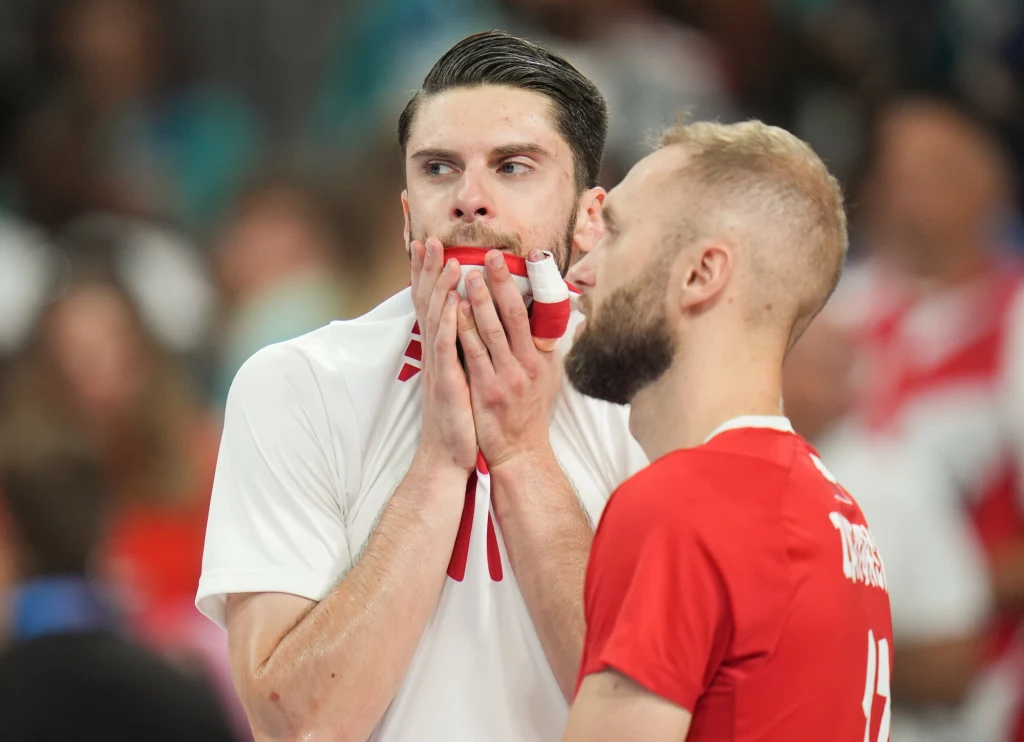Aleksander Śliwka i Paweł Zatorski Dwóch mężczyzn w sportowych strojach, jeden w białej koszulce z dłonią zakrywającą usta i wyrazem skupienia na twarzy, drugi w czerwonej koszulce z numerem 14 na plecach, w tle rozmazane postacie kibiców.