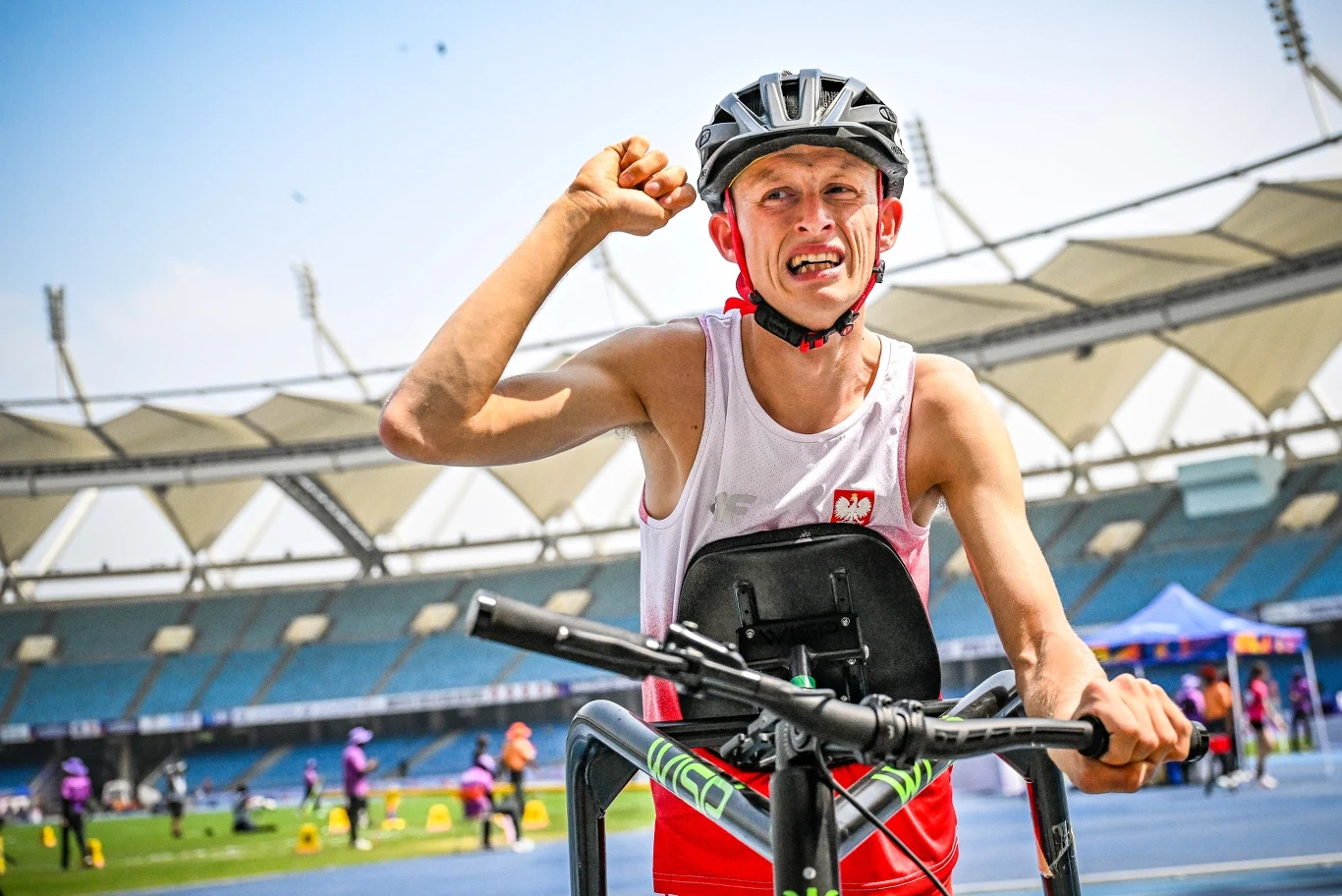 Artur Krzyżek Sportowiec w koszulce z polskim herbem i kasku rowerowym, uśmiechnięty, podczas wyścigu na stadionie lekkoatletycznym, unosi w górę zaciśniętą pięść w geście triumfu.