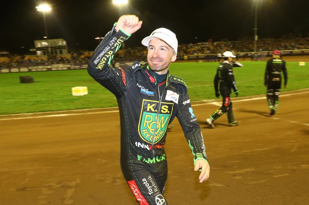 Żużlowiec ubrany w profesjonalny strój sportowy przechadza się po torze żużlowym podczas nocnych zawodów, w tle widoczny tłum kibiców oraz jasne oświetlenie stadionu.