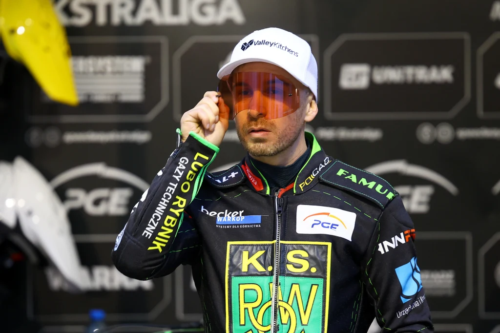 Zawodnik sportów motorowych w profesjonalnym kombinezonie z logotypami sponsorów oraz białą czapką i pomarańczowymi okularami znajduje się na tle ściany z banerami reklamowymi.