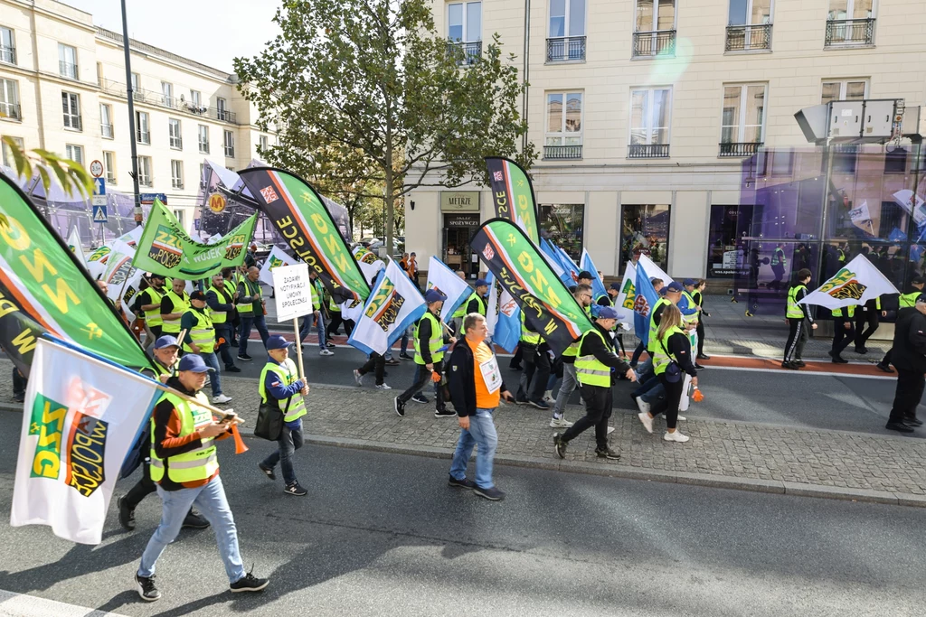 Grupa osób ubranych w kamizelki odblaskowe maszeruje ulicą z flagami i transparentami, w tle widać budynki mieszkalne oraz drzewo, atmosfera jest spokojna i zorganizowana.