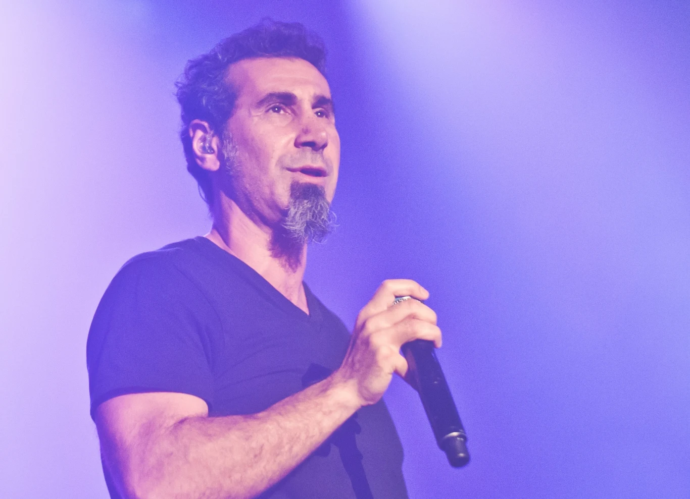 Serj Tankian Mężczyzna z krótko przyciętymi włosami i charakterystyczną bródką trzyma mikrofon, stojąc w oświetleniu scenicznym utrzymanym w fioletowo-niebieskich tonach.