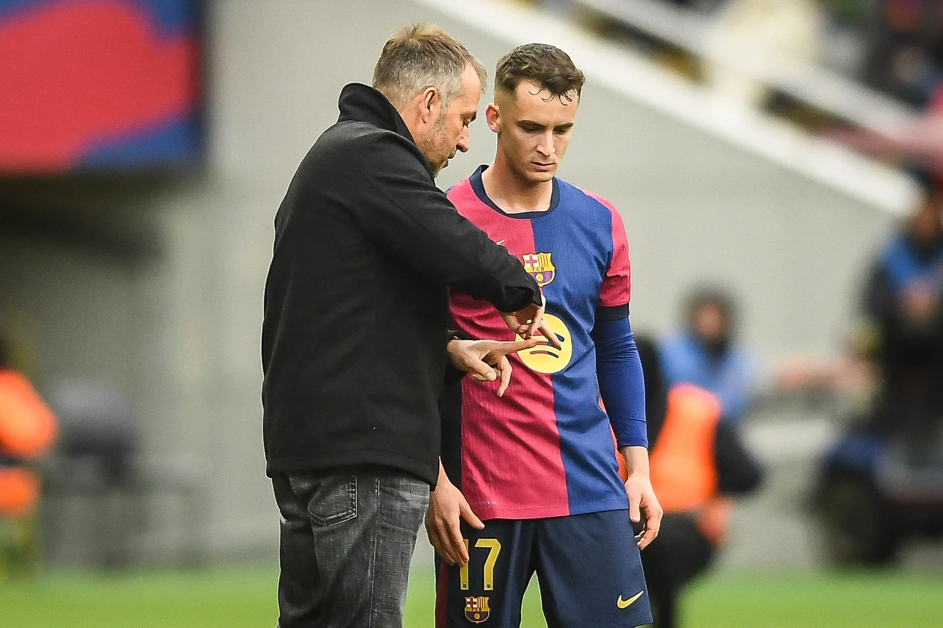 Mężczyzna w ubraniu sportowym oraz trener w kurtce, podczas rozmowy na boisku piłkarskim. Piłkarz ubrany w koszulkę klubu FC Barcelona z numerem 7, w tle niewyraźni zawodnicy i stadion.