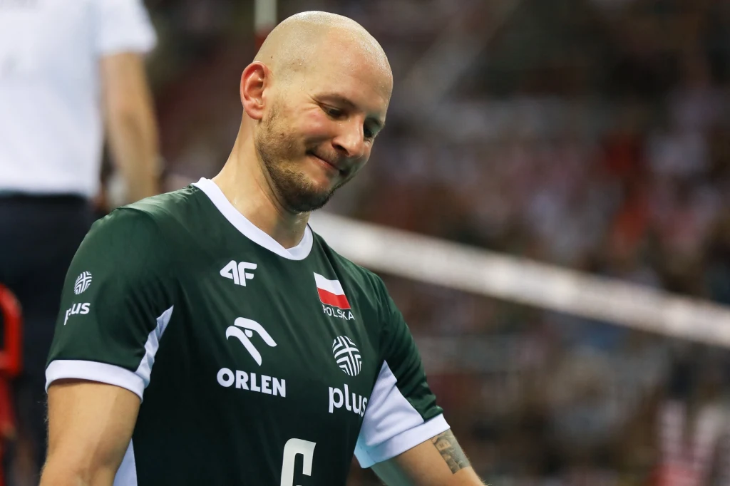 Bartosz Kurek Siatkarz w zielonej koszulce reprezentacji Polski z opuszczoną głową i lekko smutnym wyrazem twarzy, na tle rozmytej hali sportowej.