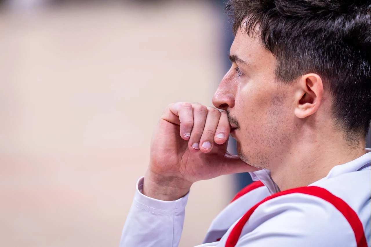 Młody mężczyzna w sportowym stroju z czerwonymi akcentami siedzi zamyślony, opierając brodę na dłoni i patrząc przed siebie.