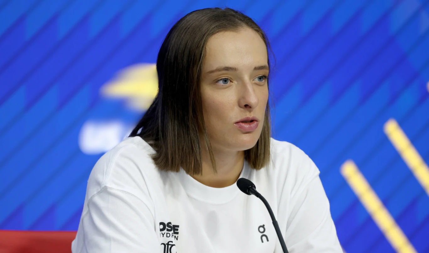 Iga Świątek Młoda kobieta o prostych, brązowych włosach udziela wywiadu lub bierze udział w konferencji prasowej, siedząc przy mikrofonie na tle niebieskiej ściany ze wzorami i logotypami.