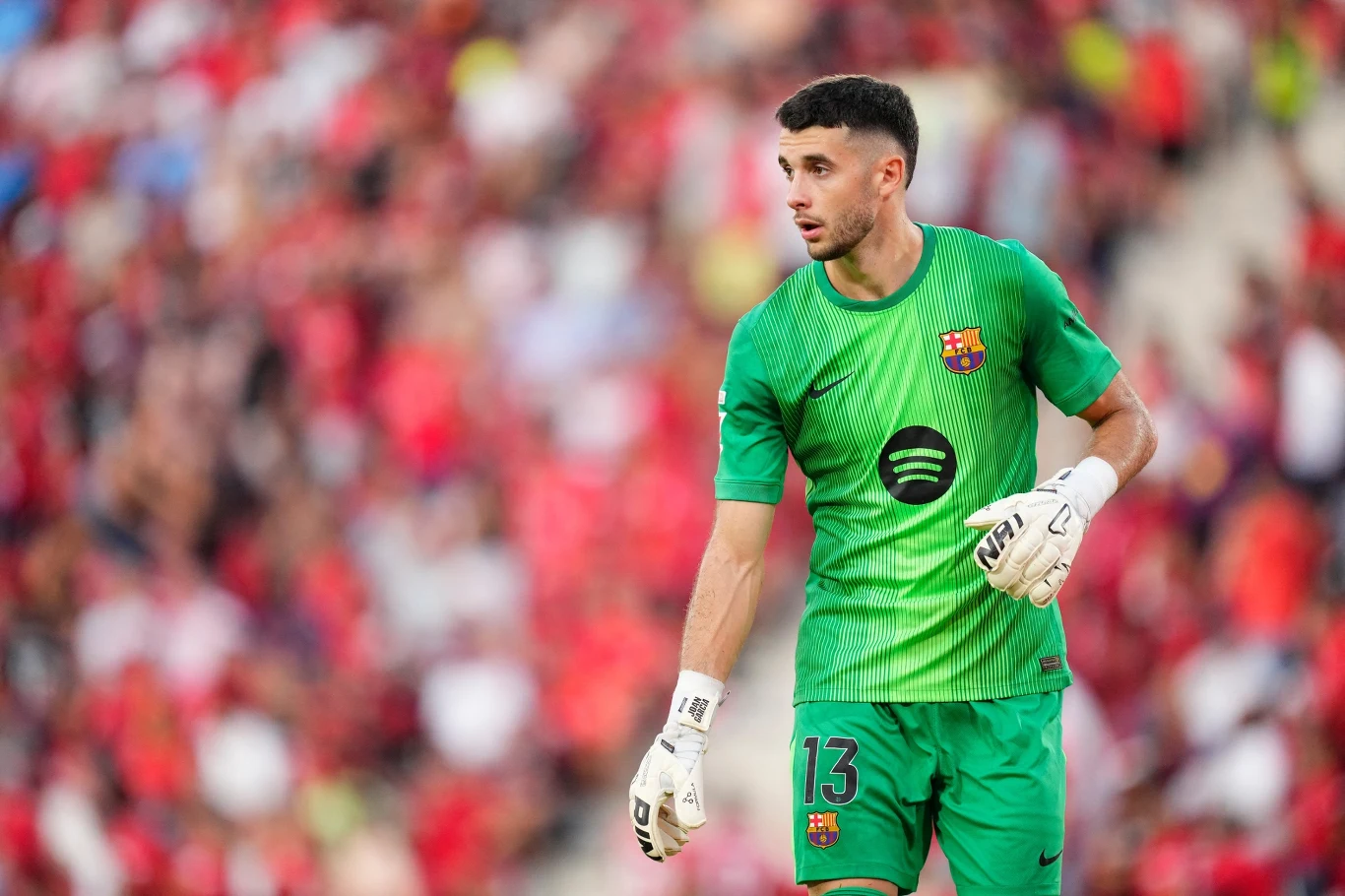 Bramkarz w zielonym stroju z logo FC Barcelony i numerem 13 na spodenkach stoi na boisku, w tle rozmyte trybuny z kibicami.