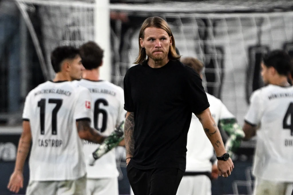 Eugen Polanski, trener Borussii Moenchengladbach Mężczyzna z długimi blond włosami w czarnym t-shircie stoi na murawie boiska, w tle kilku piłkarzy w białych koszulkach z widocznymi numerami i nazwiskami, atmosfera sportowego wydarzenia.