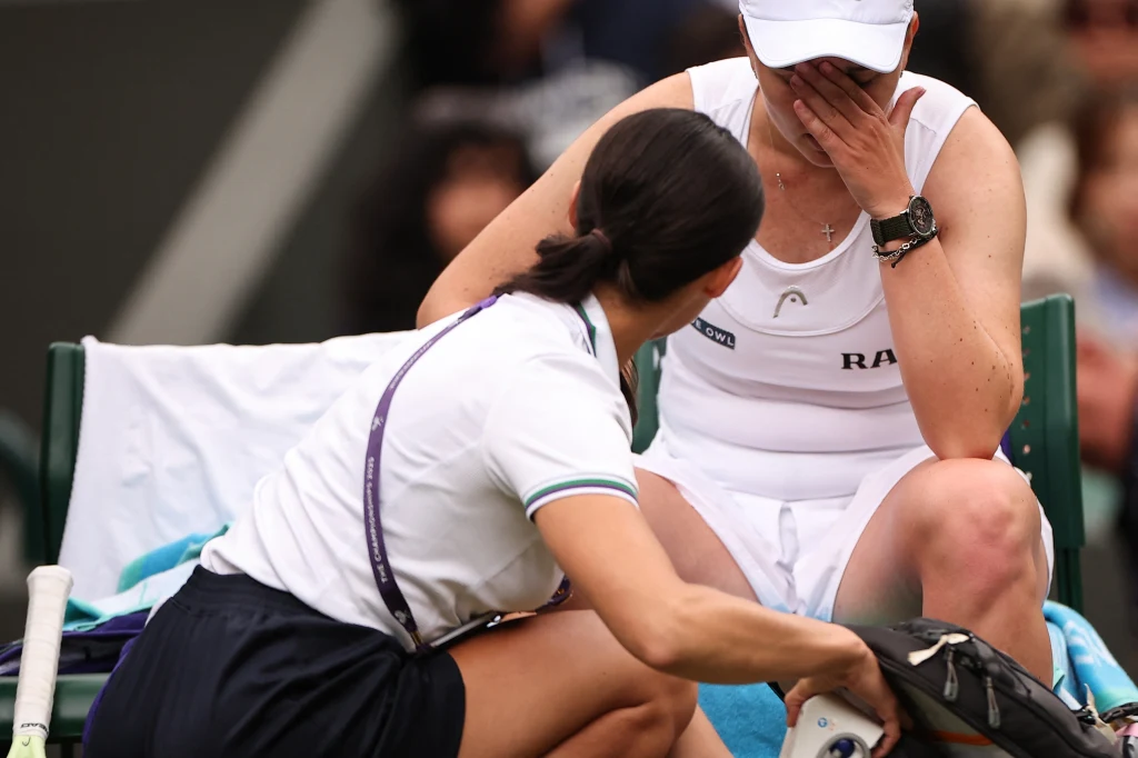 Barbora Krejcikova nie po raz pierwszy boryka się z kłopotami zdrowotnymi Zawodniczka siedząca na ławce podczas meczu tenisa zakrywa twarz dłonią, obok niej kuca fizjoterapeutka udzielająca pomocy medycznej, widoczne sportowe stroje i sprzęt.