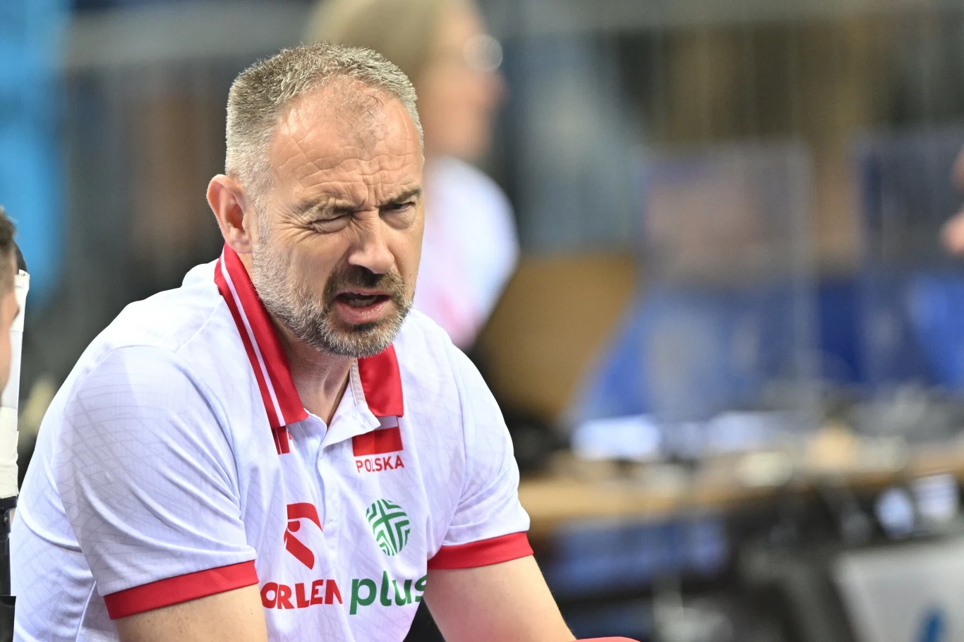 Nikola Grbić Mężczyzna w białej koszulce z czerwonymi akcentami i napisem Orlen Polska siedzi w hali sportowej, marszczy twarz i mruży jedno oko, na jego twarzy widoczna jest silna emocja.