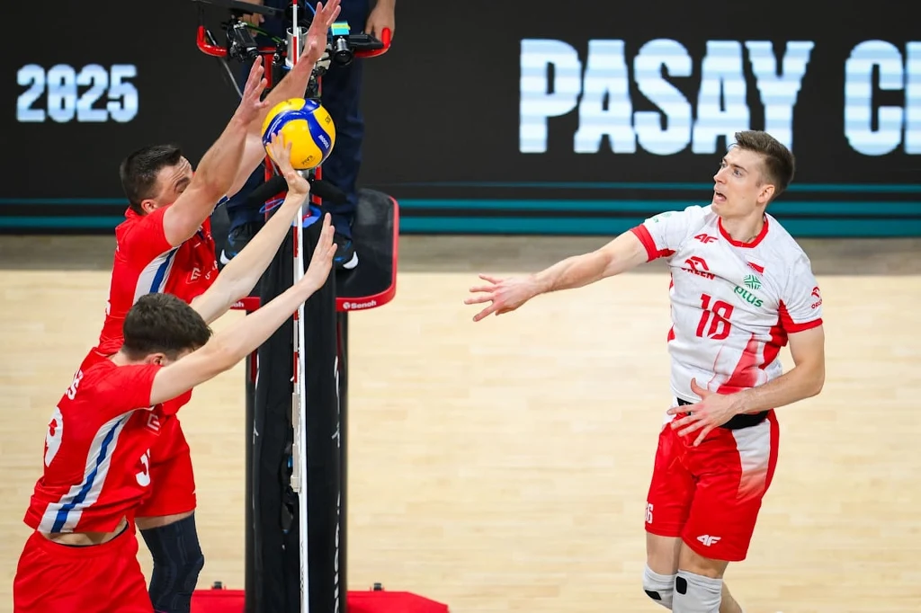 Kamil Semeniuk w meczu Polska - Czechy Siatkarz w biało-czerwonym stroju wykonuje atak na piłkę ponad siatką, po drugiej stronie dwaj zawodnicy w czerwonych strojach próbują zablokować piłkę, dynamiczna akcja podczas meczu siatkówki.