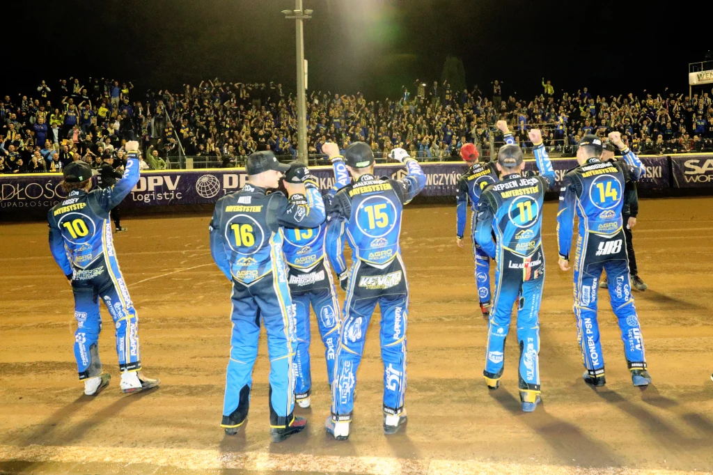 zawodnicy GKM-u Grudziądz cieszą się z kibicami Grupa żużlowców ubranych w niebieskie kombinezony z numerami na plecach celebruje sukces na torze, podnosząc ręce w kierunku tłumu kibiców zgromadzonych na trybunach.
