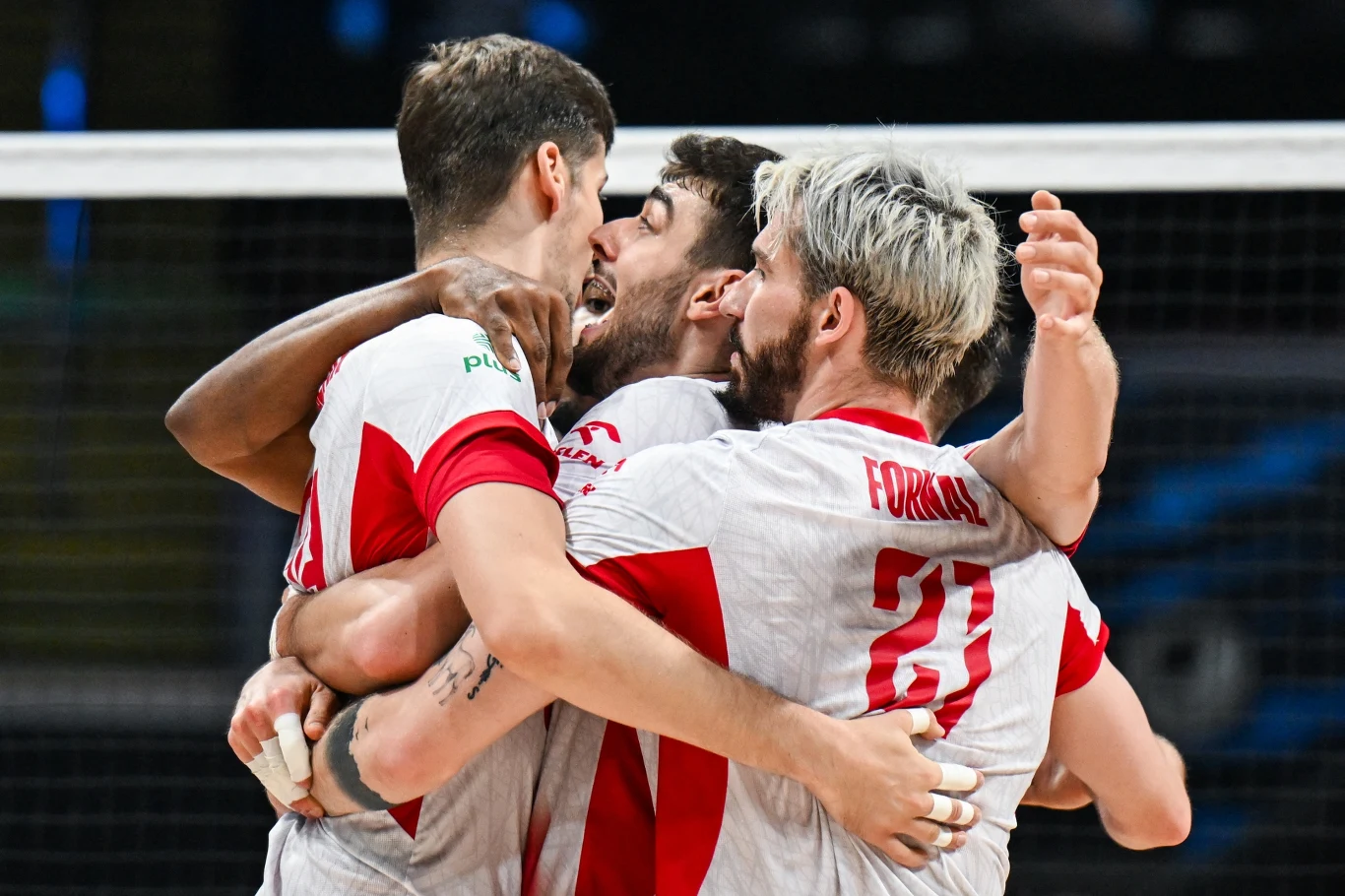 Grupa siatkarzy w biało-czerwonych strojach obejmuje się i wyraża radość po zdobytym punkcie, dominują gesty triumfu i sportowej jedności, w tle widoczna siatka.