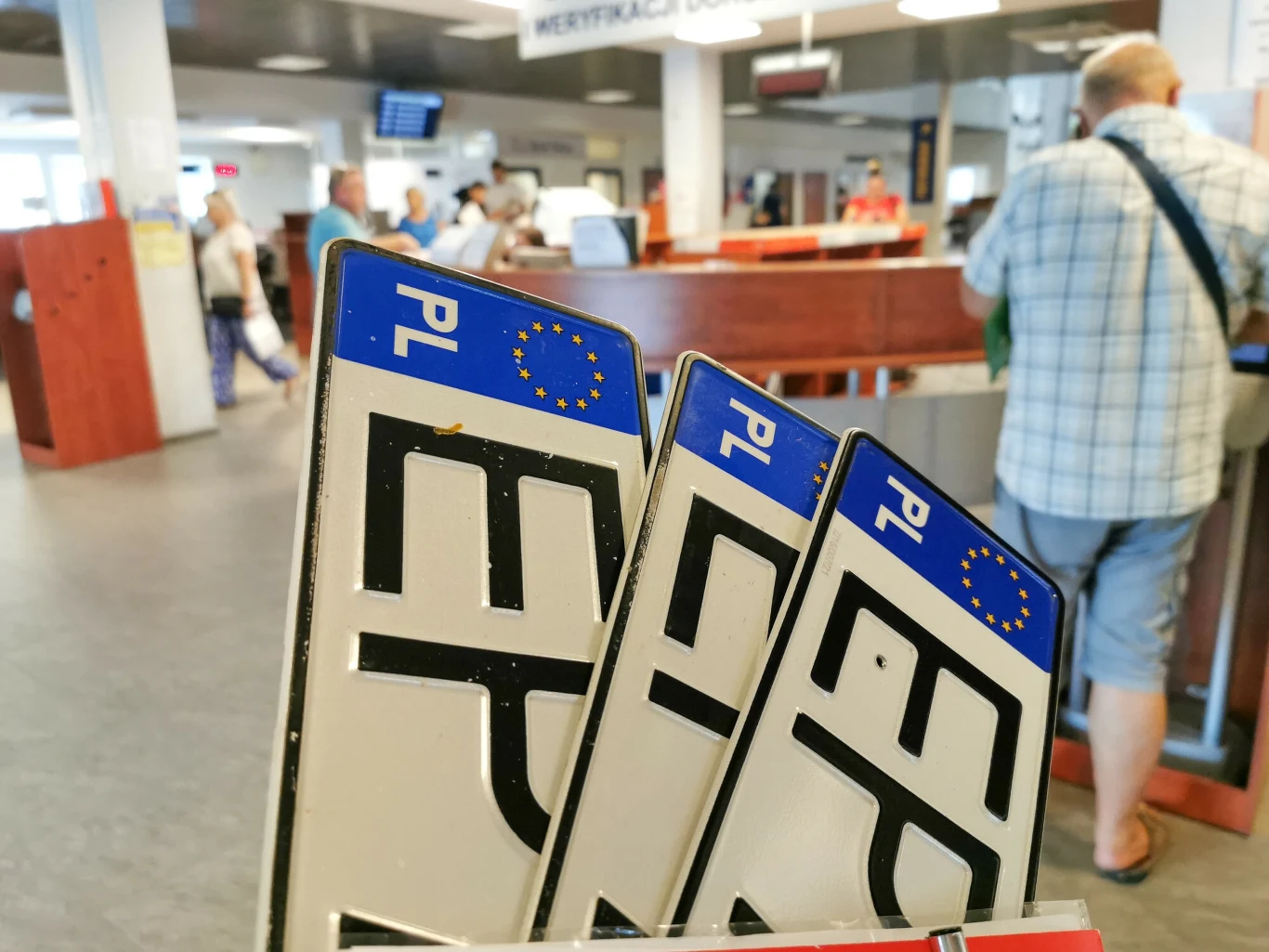 Będą ułatwienia dla osób rejestrujących samochody Zbliżenie na trzy polskie tablice rejestracyjne umieszczone na pierwszym planie, w tle wnętrze urzędu z osobami oczekującymi w kolejce do stanowisk obsługi.
