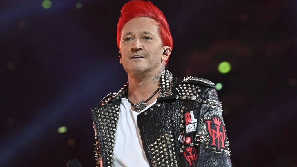 Mężczyzna z czerwonymi włosami ubrany w czarną skórzaną kurtkę z ćwiekami i naszywkami, stoi na scenie podczas występu muzycznego przy mikrofonie, w tle rozmyte kolorowe światła.