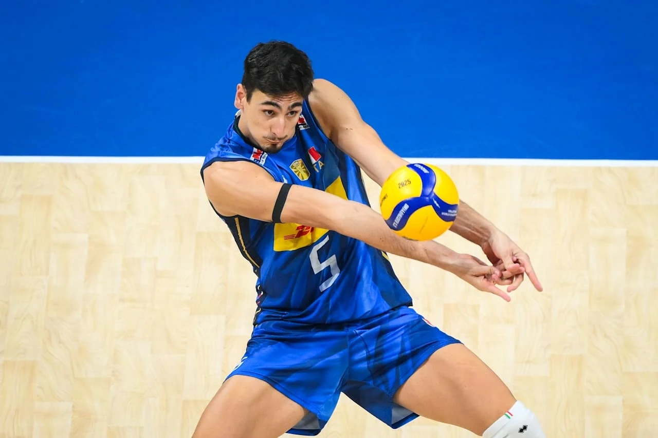 Siatkarz w niebieskim stroju wykonujący odbiór piłki podczas meczu, dynamiczna poza, intensywne skupienie na twarzy sportowca, piłka w górnej części przy jego rękach, jasne boisko sportowe jako tło.