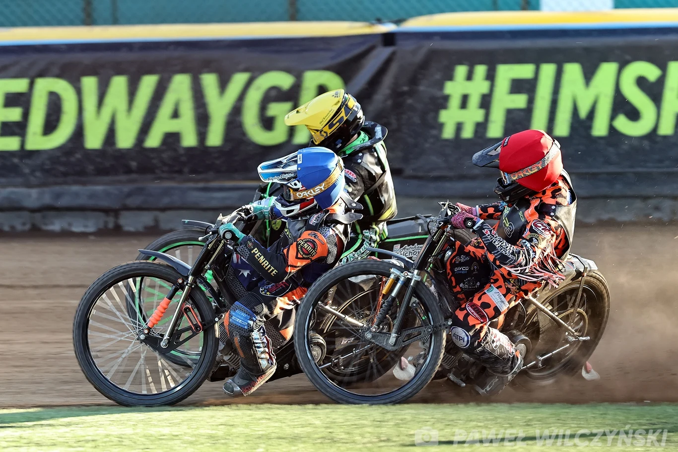 Trzech żużlowców ściga się na torze, zbliżając się do zakrętu, każdy z nich w kolorowym kombinezonie i kasku, na tle ogrodzenia z widocznymi banerami reklamowymi.