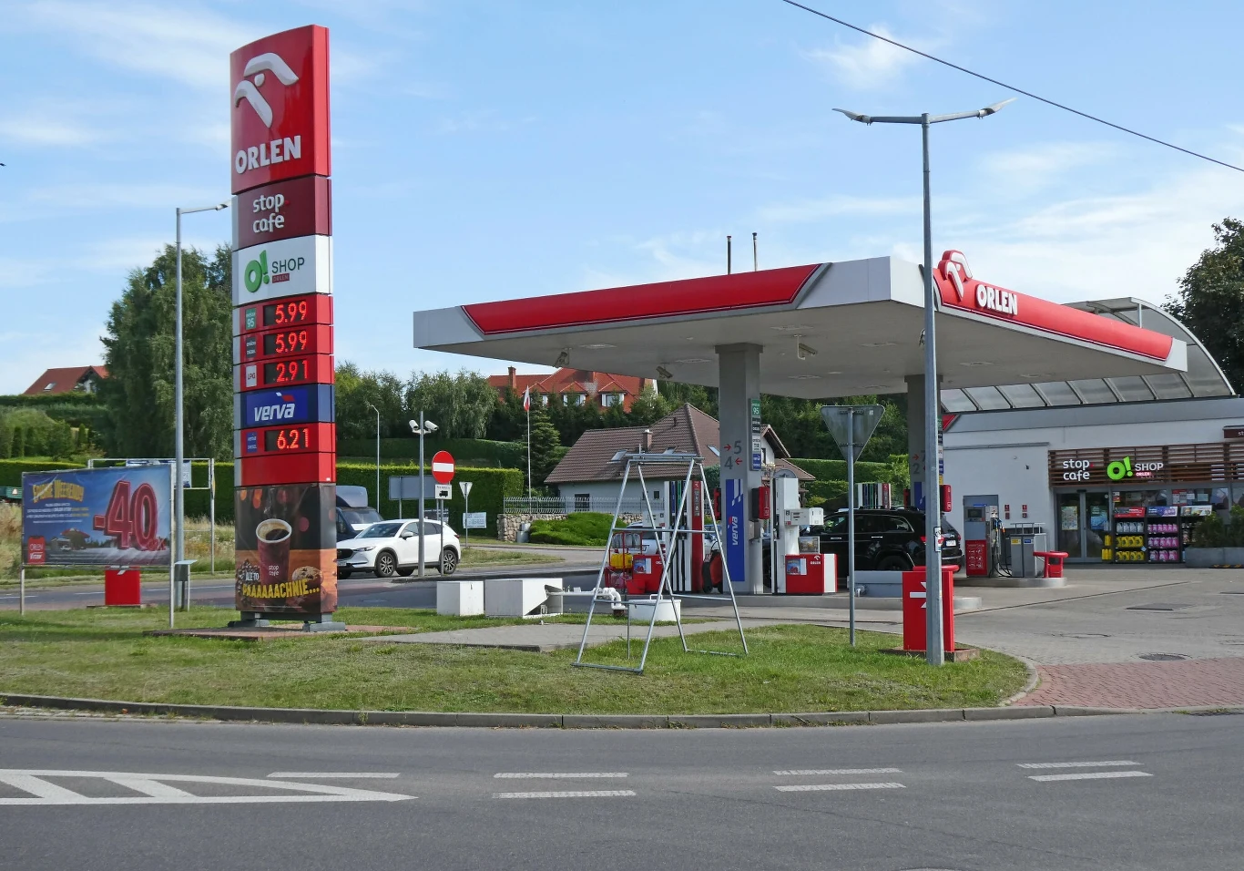 Kompresor znajdziemy na każdej stacji, więc co pewien czas, przy okazji tankowania warto sprawdzić ciśnienie w kołach Stacja benzynowa Orlen z zadaszonym stanowiskiem dla samochodów, widocznymi dystrybutorami paliwa i tablicą z aktualnymi cenami paliw. W tle sklep i reklama promocyjna, po lewej stronie wjazd z drogą asfaltową oraz otoczenie z zielenią i zabudową miesz...