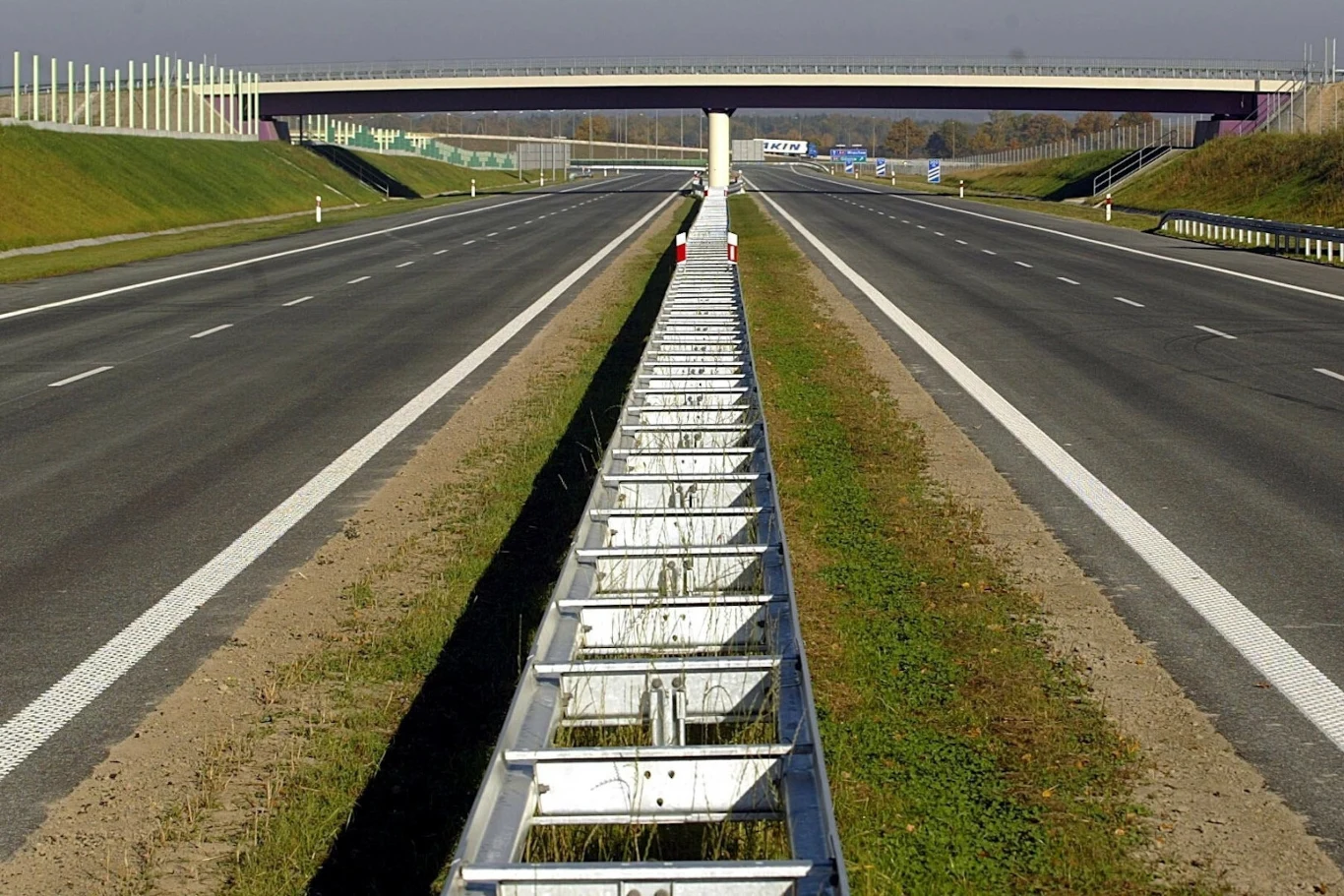 Bariery energochłonne również mogą być smart Szeroka autostrada z dwoma jezdniami oddzielonymi metalowymi barierkami pośrodku, a w tle widoczny most nad jezdniami, okolona nasypami trawiastymi oraz ekranami dźwiękochłonnymi.