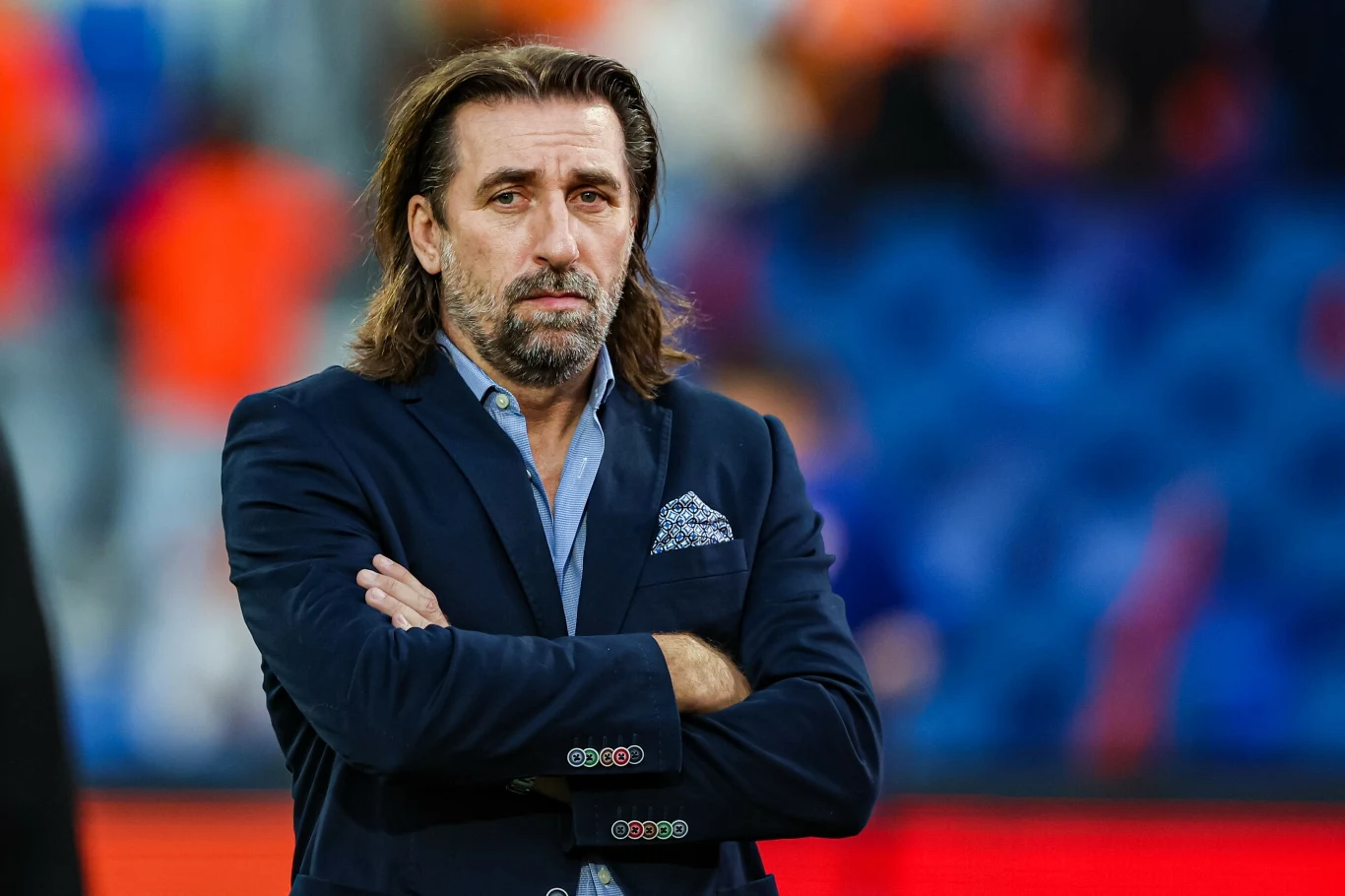 Mężczyzna w średnim wieku w eleganckiej marynarce z poszetką, stoi z założonymi rękami na tle rozmytego stadionu, patrzy poważnie w stronę obiektywu.