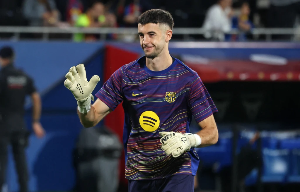 Bramkarz w sportowej koszulce z rękawicami do piłki nożnej, znajduje się na stadionie, w tle widoczne osoby oraz trybuny.