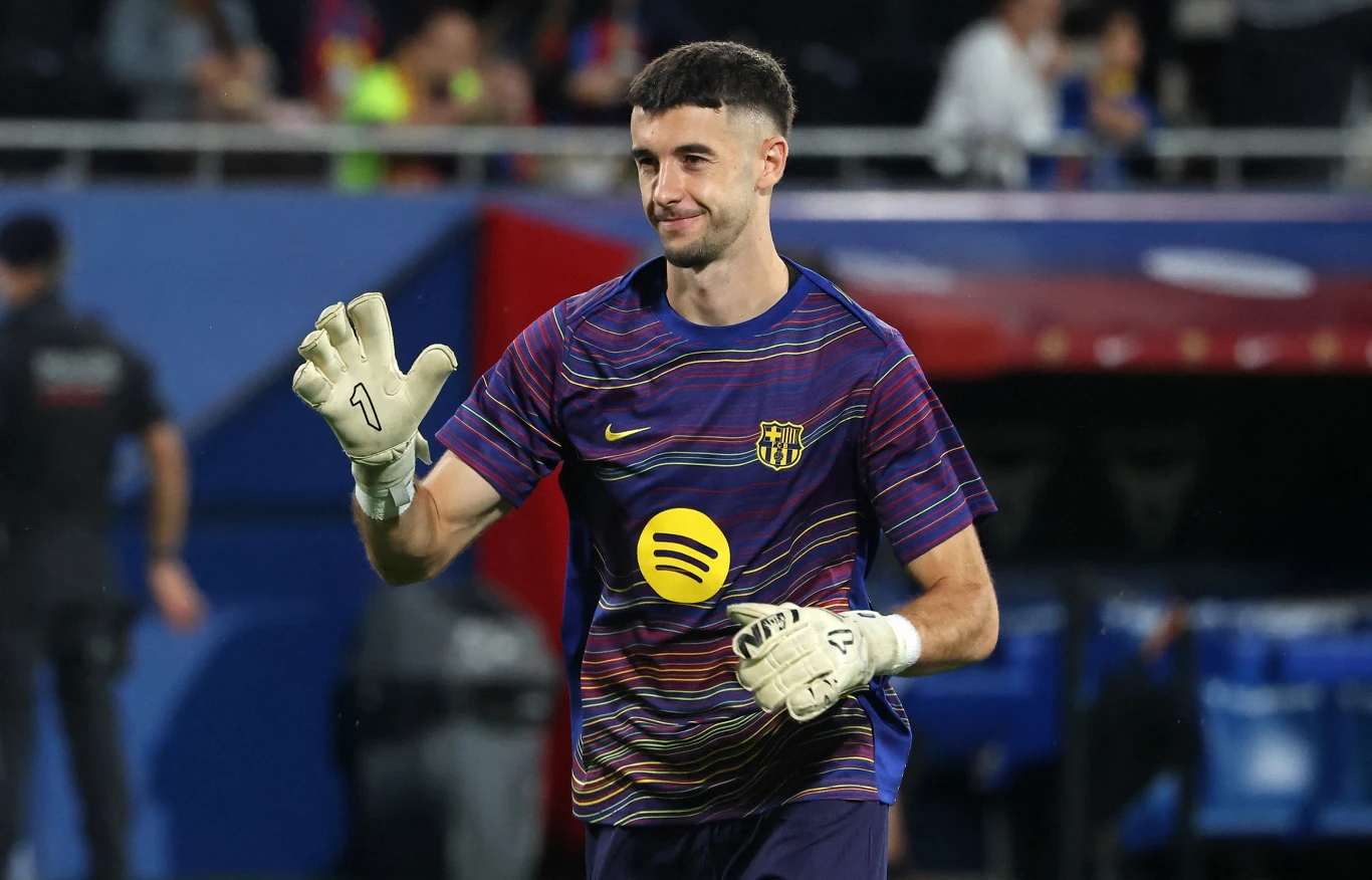 Bramkarz w sportowej koszulce z rękawicami do piłki nożnej, znajduje się na stadionie, w tle widoczne osoby oraz trybuny.