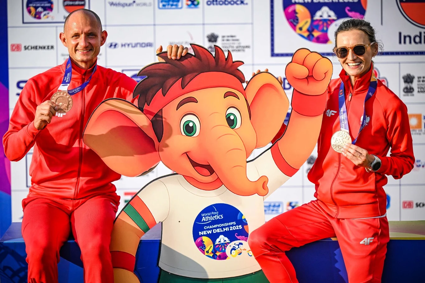 Dwóch mężczyzn w czerwonych strojach sportowych pozuje z medalami obok kolorowej tekturowej postaci słonia, na tle ściany z logotypami sponsorów oraz napisem wskazującym na wydarzenie sportowe w New Delhi w 2023 roku.