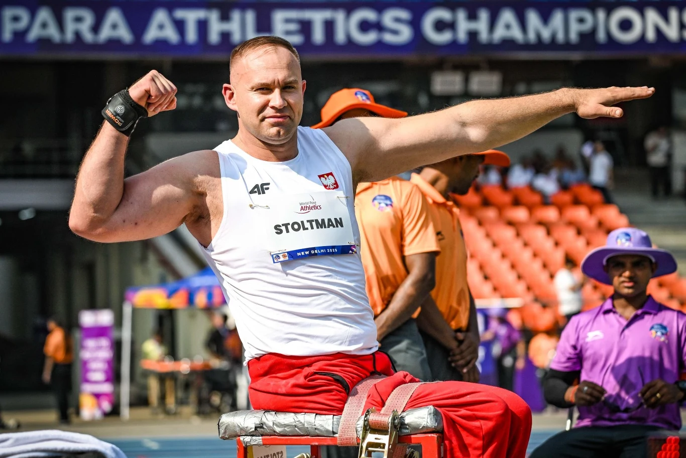 Sportowiec z niepełnosprawnością siedzi na specjalnym sprzęcie na stadionie i prezentuje muskulaturę, ubrany w biały podkoszulek z napisem Polski Związek Paraolimpijski i nazwiskiem Stoltman, w tle widoczni są sędziowie oraz puste pomarańczowe trybuny.
