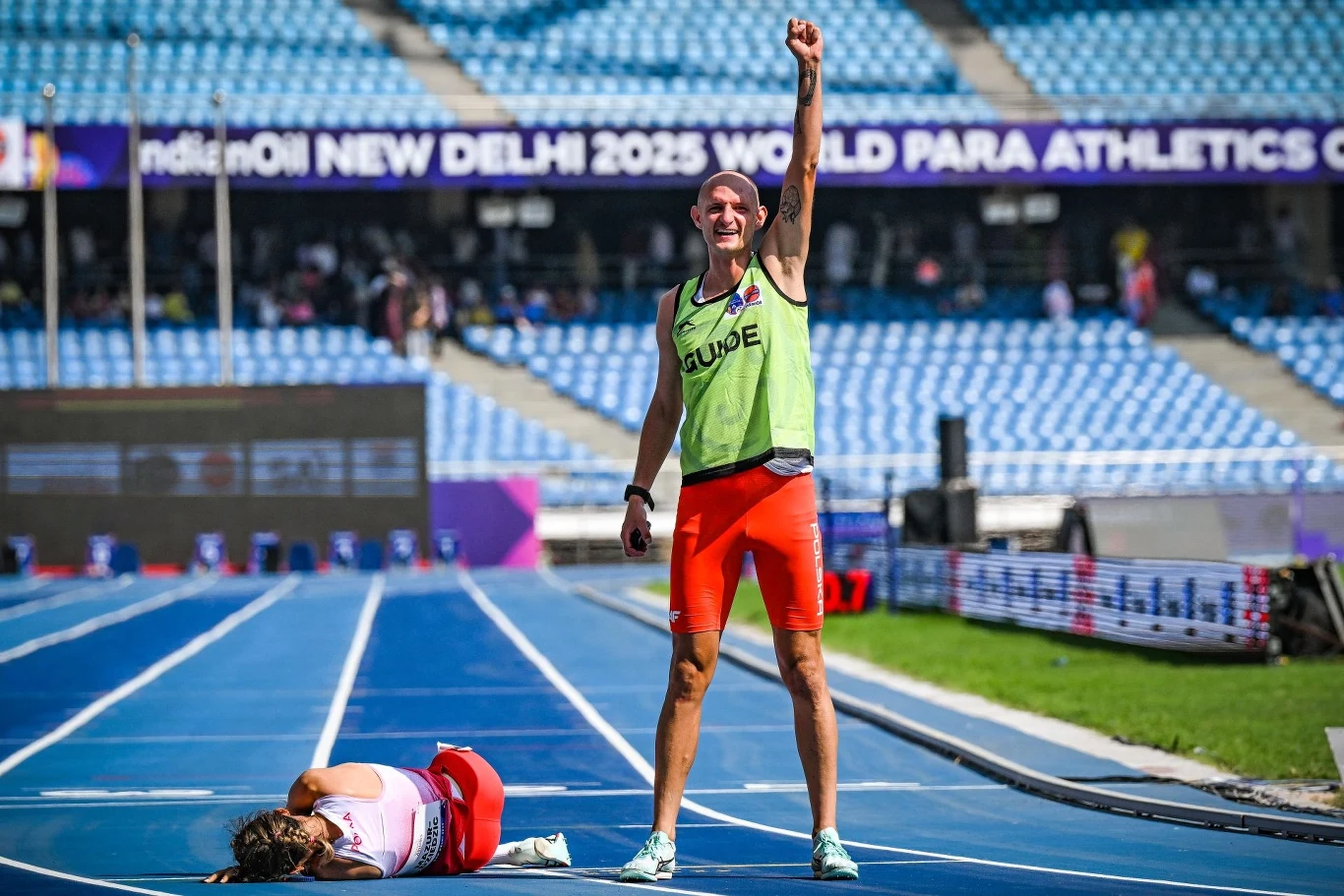 Zwycięski sportowiec z uniesioną ręką świętuje sukces na bieżni tuż obok osoby leżącej na torze, która wygląda na wyczerpaną lub pokonaną, w tle pusty stadion i baner zawodów paraatletycznych.