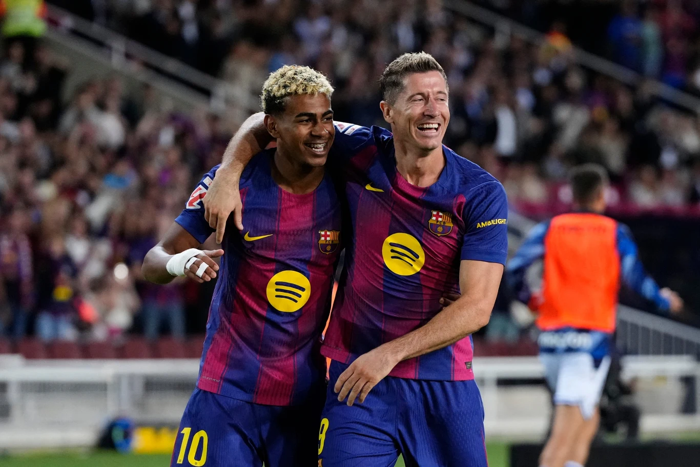 Dwóch piłkarzy drużyny FC Barcelona przytulających się i uśmiechających na tle stadionu podczas meczu, obaj ubrani w granatowo-bordowe stroje z widocznymi logotypami.