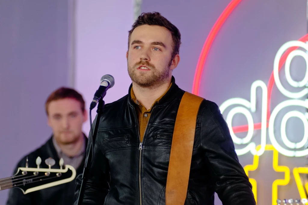 Mężczyzna w skórzanej kurtce stoi przed mikrofonem z gitarą na ramieniu, za nim widać drugiego muzyka oraz fragment jasnego neonowego napisu na ścianie.