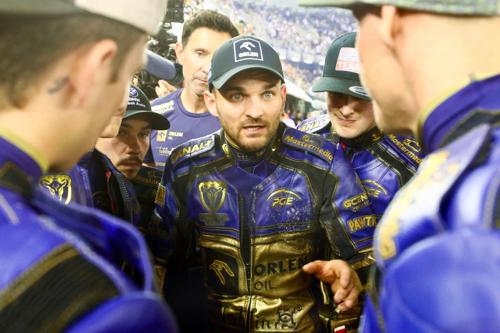 Grupa żużlowców w niebiesko-złotych kombinezonach z logo sponsorów omawia strategię podczas zawodów, centralnie ujęty zawodnik dynamicznie gestykuluje i skupia wzrok na rozmówcach.