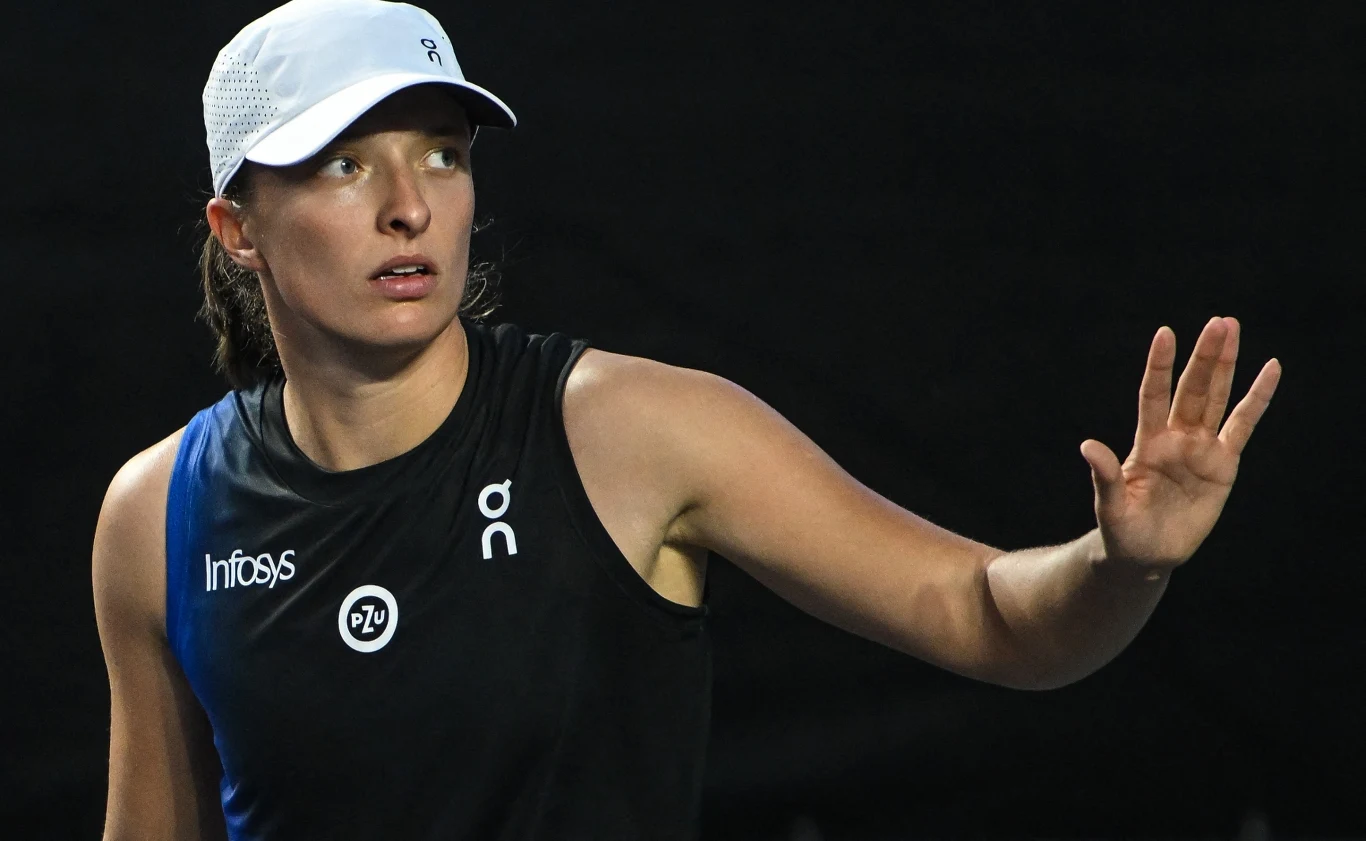 Sportowiec w sportowym stroju i białej czapce wykonuje gest dłonią, skupiona twarz wskazuje na koncentrację, tło jest ciemne, co podkreśla sylwetkę.