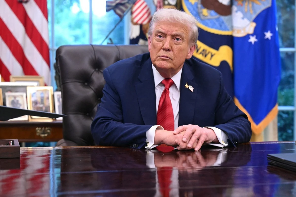 Donald Trump Mężczyzna w garniturze i czerwonym krawacie siedzi przy dużym, drewnianym biurku w eleganckim gabinecie, w tle widoczne symbole państwowe oraz zdjęcia rodzinne.