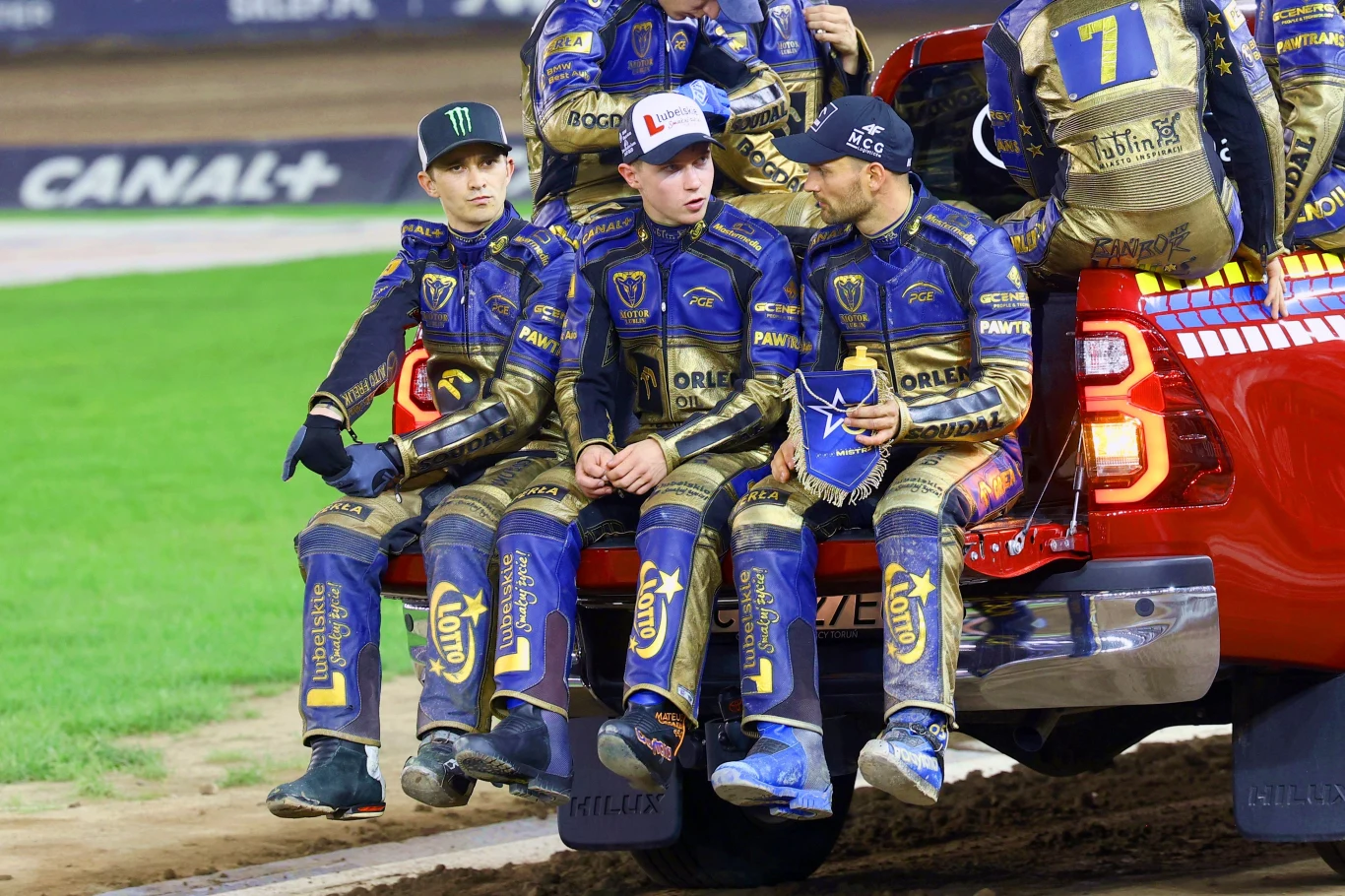 Zawodnicy w kombinezonach motorowych siedzący na tylnej części czerwonego pickupa na torze żużlowym, rozmawiający ze sobą, w tle arena sportowa.