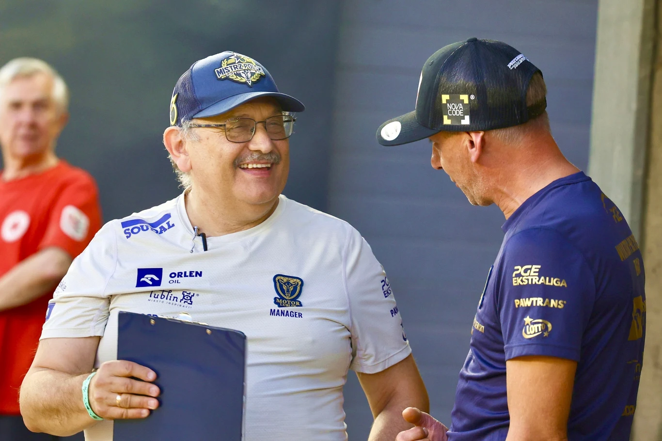 Dwóch mężczyzn w sportowych koszulkach rozmawia ze sobą, jeden z nich trzyma w ręku clipboard, obaj uśmiechnięci, w tle rozmazana postać w czerwonej koszulce.