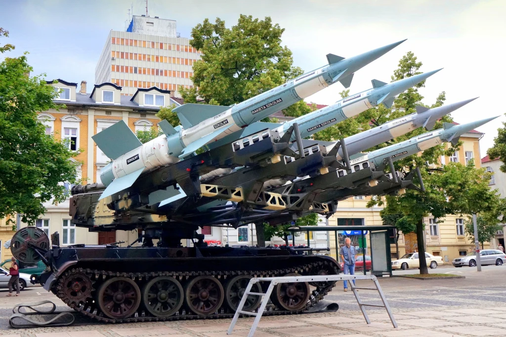 Wyrzutnia rakietowa SAM z czterema dużymi pociskami umieszczona na gąsienicowym podwoziu, ustawiona na placu miejskim, w otoczeniu zielonych drzew oraz budynków mieszkalnych i biurowych.