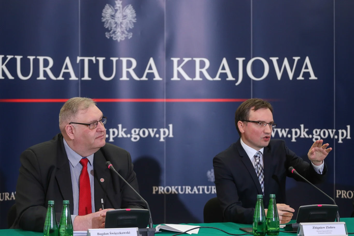 Dwóch mężczyzn w garniturach siedzi przy stole przykrytym zielonym obrusem, przed nimi stoją tabliczki z imionami oraz mikrofony, za nimi znajduje się tło z napisem Prokuratura Krajowa i adresem internetowym, na stole stoją zielone butelki z wodą.
