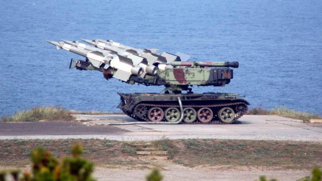 Wyrzutnia rakietowa zamontowana na podwoziu gąsienicowym ustawiona na utwardzonym placu blisko wybrzeża z widocznym morzem w tle