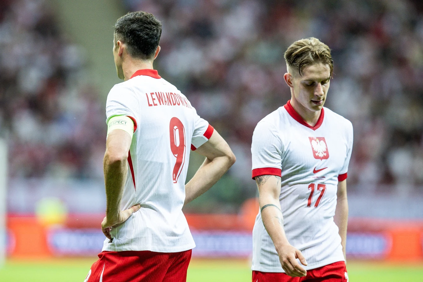 Robert Lewandowski i Michał Skóraś w reprezentacji Polski Dwóch piłkarzy w białych koszulkach reprezentacji Polski, jeden z numerem 9 i nazwiskiem Lewandowski na plecach, drugi z numerem 17, stojący na boisku, wyrażający zamyślenie lub rozczarowanie, widoczne trybuny i kibice w tle.