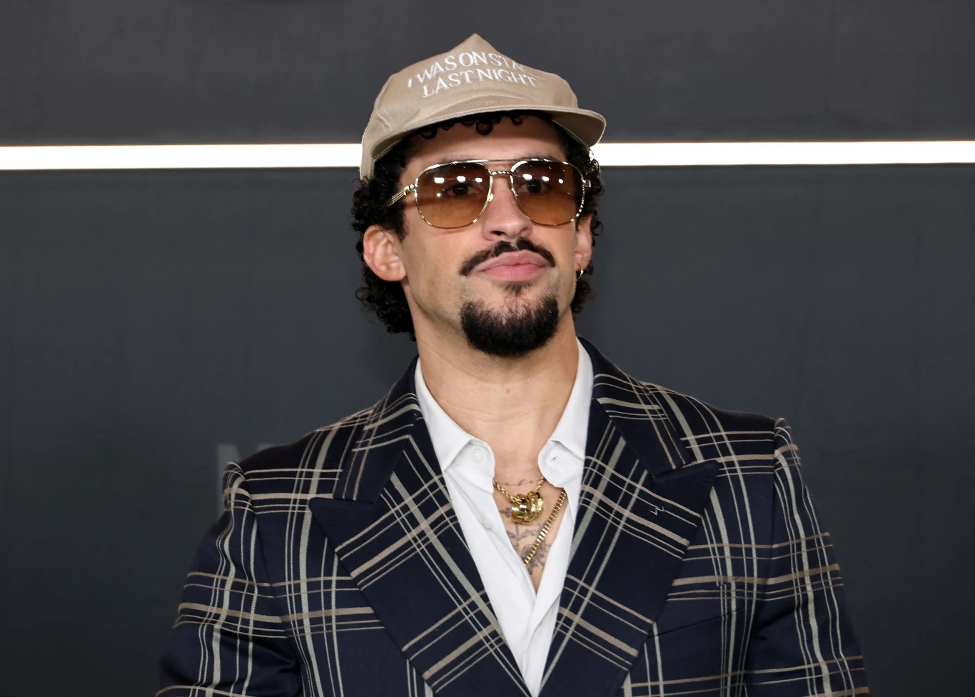 Mężczyzna w okularach przeciwsłonecznych i beżowej czapce z daszkiem, ubrany w wzorzystą marynarkę i biały podkoszulek, z naszyjnikami na szyi, tło jednolite, ciemne.