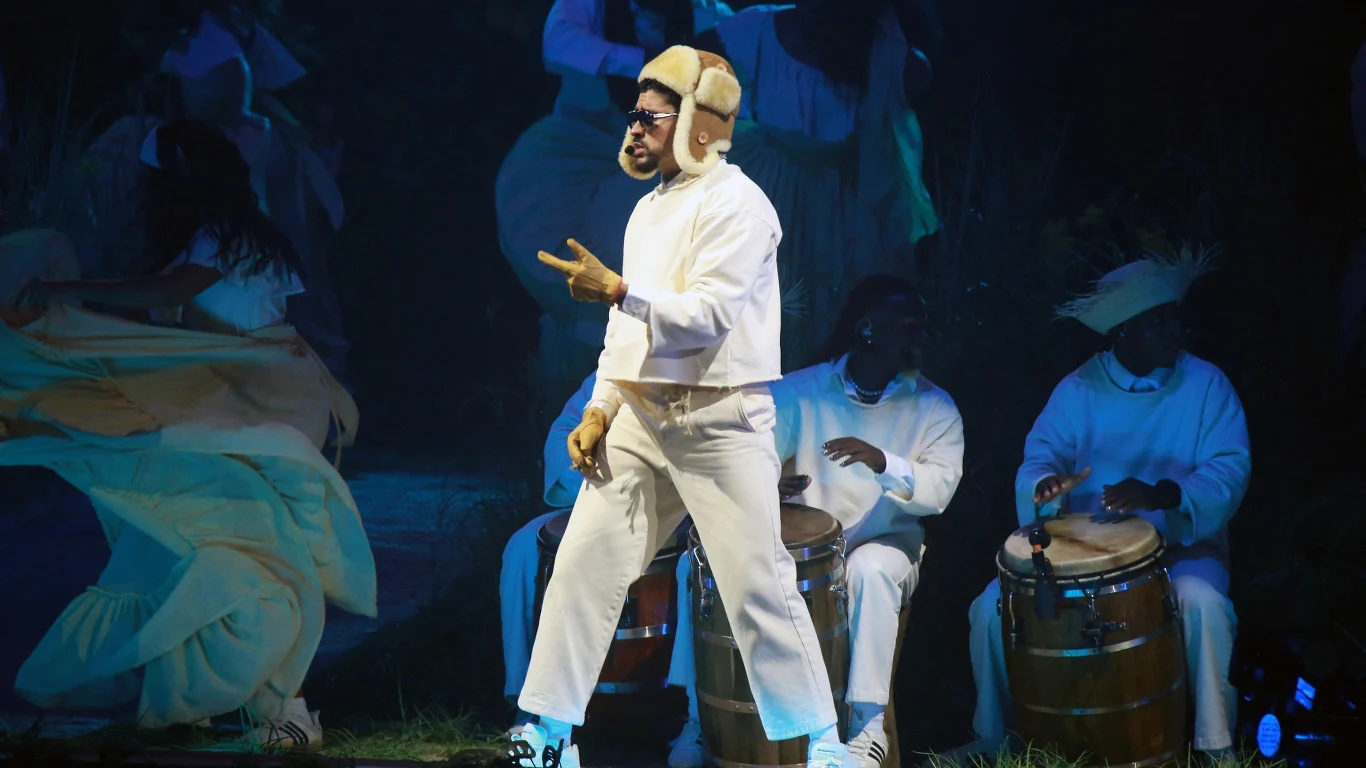 Mężczyzna w jasnym ubraniu oraz czapce pilotce śpiewa lub rapuje na scenie, w tle muzycy grający na bębnach również ubrani na biało, światła sceniczne podkreślają teatralny nastrój.