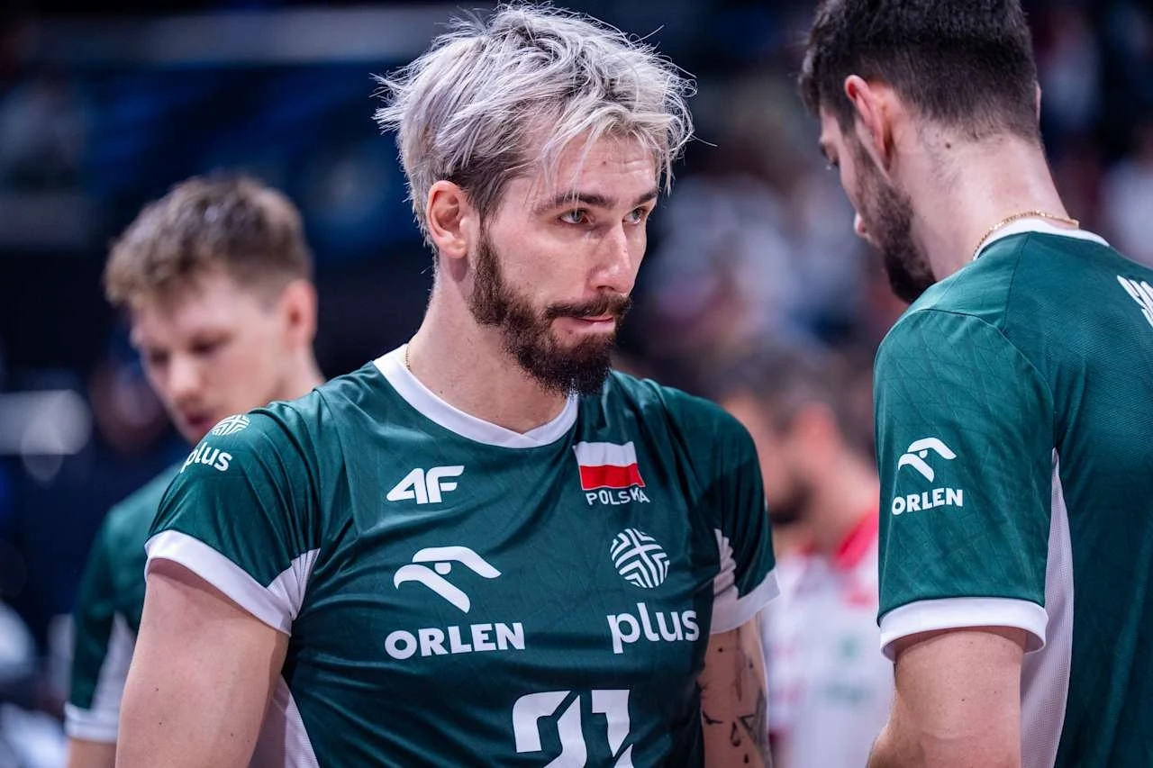 Zawodnik drużyny siatkarskiej ubrany w zielony strój z polską flagą i logo sponsorów, skupiony podczas rozmowy z kolegą z drużyny, w tle rozmazani inni zawodnicy.