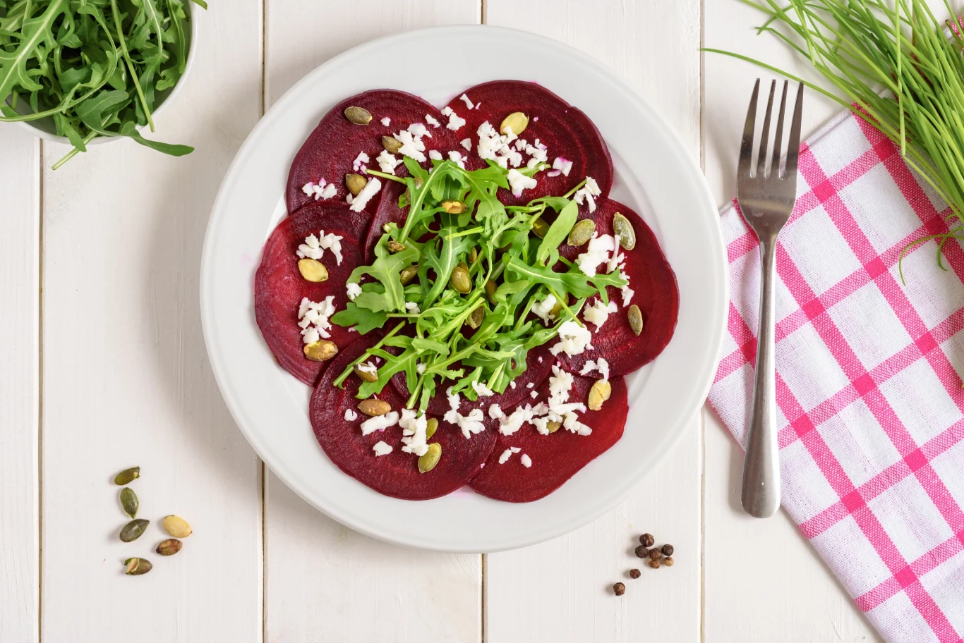Carpaccio buraczane z miodem - przepis Ewy Wachowicz Plasterki buraka ułożone w okrąg na talerzu, posypane pokruszoną fetą, pestkami dyni i świeżą rukolą, obok widoczne zioła oraz sztućce na białym stole z różowo-białą serwetką.