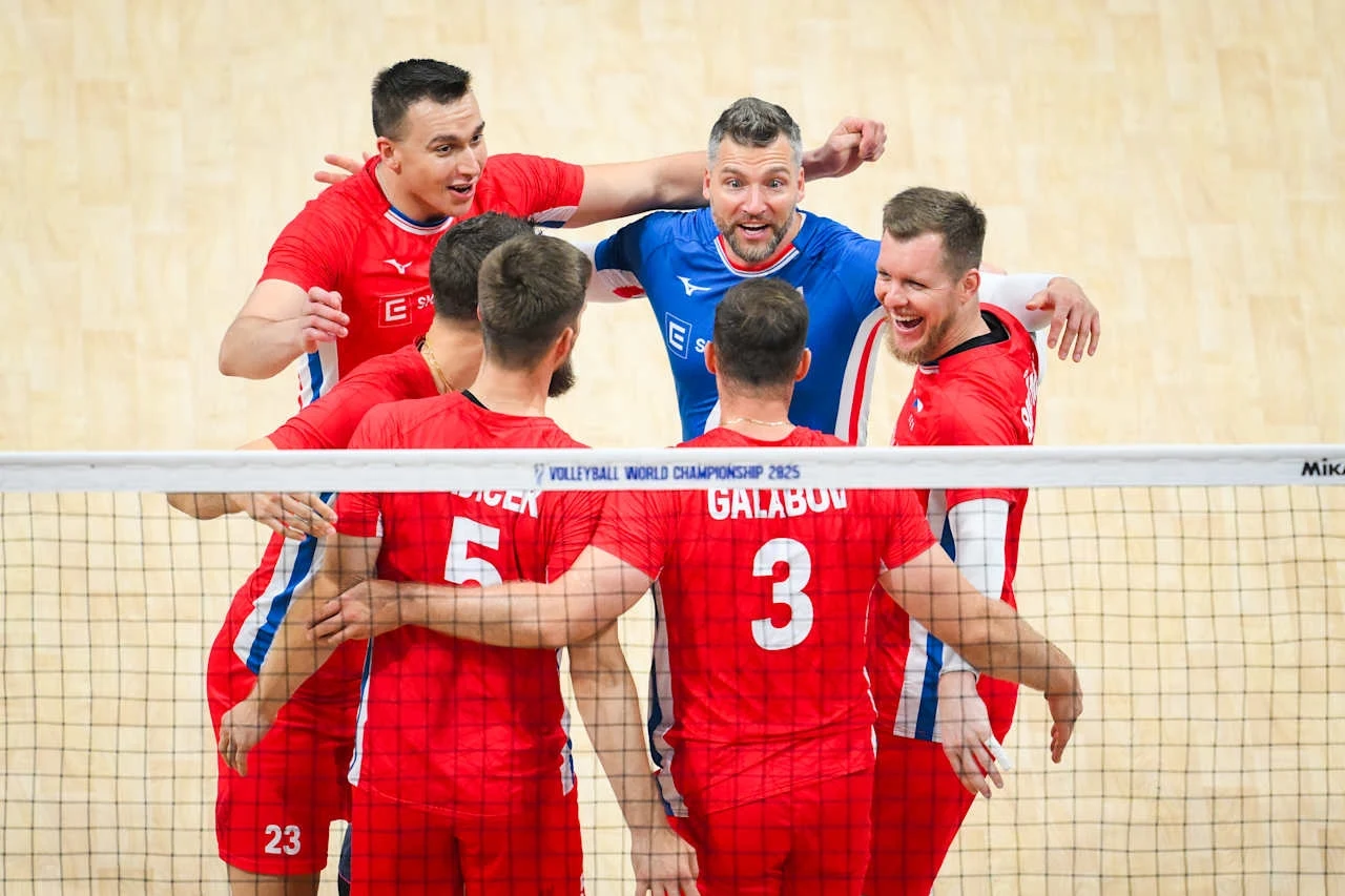 Siatkarze reprezentacji Czech, z nr 5 Adam Zajicek Grupa sześciu siatkarzy ubranych w czerwone stroje świętuje razem zdobyty punkt w trakcie meczu siatkówki, stojąc w kręgu za siatką na jasnym parkiecie.