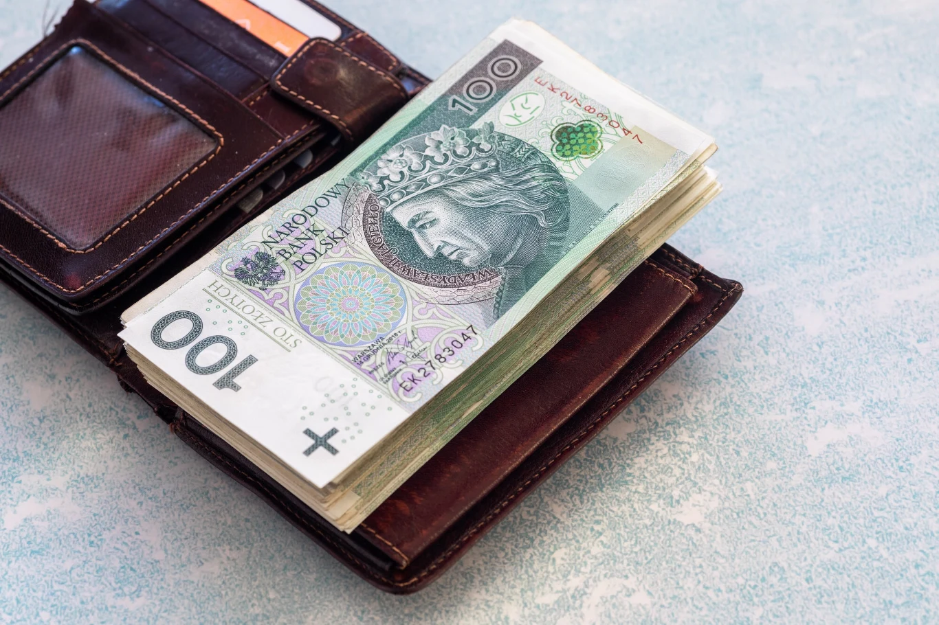 GUS podał dane o rozkładzie wynagrodzeń w gospodarce narodowej w kwietniu 2025 r. Skórzany portfel na stole z dużym plikiem stuzłotowych banknotów polskich oraz widocznymi przegródkami na karty.